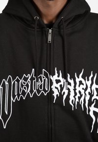 Sudadera con cremallera negra y cremallera plateada, que presenta un gran texto en estilo de llama blanca en la parte delantera y cordones en la capucha.