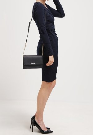 Nainen mustassa pitkähihaisessa mekossa, joka käyttää mustia korkokenkiä ja kantaa mustaa nahkainen olkalaukkua kultaketjulla.