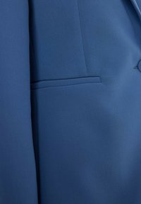 Niebieska marynarka z tkaniny o strukturalnym designie, z małą, ukośną kieszenią i gładką fakturą. Brak widocznych elementów metalowych lub akcentów.