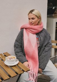 Pink ombre scarf made of soft material with fringed ends, worn over a gray quilted jacket. Coffee in a white cup on a wooden table.