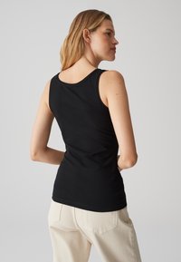 Femme aux cheveux blonds portant un top noir sans manches et un pantalon beige clair, debout, mains dans le dos, dos tourné.