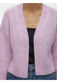 Cardigan lavorato a maglia in morbida lana rosa, con scollatura a V profonda, bordi a coste e vestibilità rilassata. Indossato sopra una maglietta nera con jeans blu.