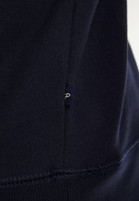 Marineblauwe katoenstof met een gestructureerde afwerking. Bevat een klein, genaaid label met witte letters en contrasterende stiksels.