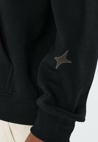 Sudadera negra hecha de una tela suave con puños acanalados. Presenta un acento brillante en forma de estrella en la manga izquierda. Diseño simple, sin patrones.
