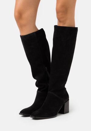 Bottes hautes en daim noir jusqu'aux genoux avec talons carrés portées sur des jambes nues, debout sur une surface blanche.
