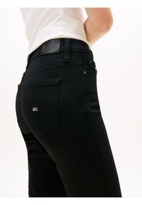 Person wearing black high-waisted jeans with a small logo on the back pocket, paired with a white ribbed top.