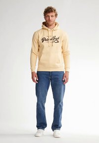 Žlutá mikina s kapucí s nápisem "Petrol Ind Co" hnědým tiskem, přední kapsa na břiše, kombinovaná s modrými džínami a bílými teniskami. Ležérní střih a měkká textura.