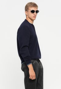 Jonge man met donkere zonnebril, gekleed in een marineblauwe ritswoller en zwarte jeans, staande opzij tegen een eenvoudige witte achtergrond.