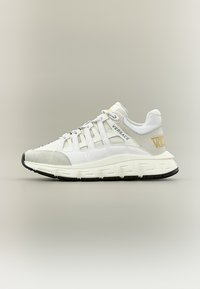 Baskets Versace blanches et gris clair avec design à lacets, semelle noire, et détail du logo "Versace" doré sur le talon et le côté.