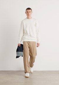 Hvit genser med grafisk detalj, beige bukser med bred ben, hvite sneakers, holder en blå og grå brettet jakke, minimalistisk bakgrunn.