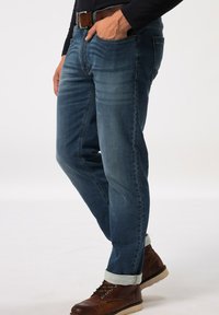 Jeans de mezclilla azul oscuro con corte ajustado, con costuras en contraste y bolsillos frontales. Los dobladillos enrollados muestran un forro azul claro; se combinan con botas marrones.