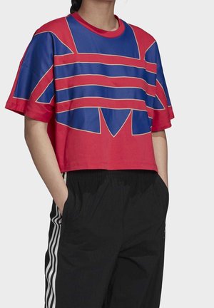 Przycięta czerwona koszulka z niebieskim wzorem graficznym na piersi, o luźnym kroju i krótkich rękawach. Połączona z czarnymi spodniami.