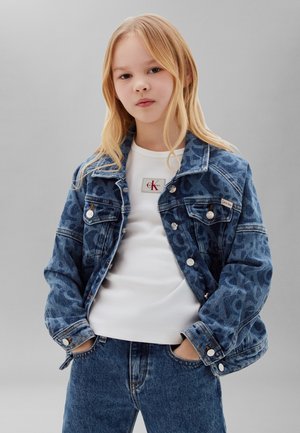 Blondes Mädchen trägt eine blaue gemusterte Jeansjacke über einem weißen Calvin Klein Shirt, Hände in den Taschen der blauen Jeans, neutraler grauer Hintergrund.