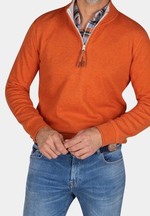 Mand iført orange kvartlynet sweater over ternet skjorte, justerer bælte på blå jeans, vist fra bryst til hofter.