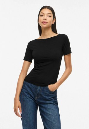 Femme aux longs cheveux foncés portant un haut noir ajusté à manches courtes et un jean bleu, posant avec une main dans la poche devant un fond uni.