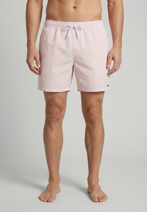 Mannelijk model draagt lichtroze gestreepte zwemshorts met wit trekkoord en klein groen krokodillenlogo op het linkerbeen, staand op blote voeten.