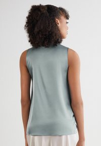 Bezramenný top v světle modrošedé barvě, vyrobený z hladké látky, s uvolněným střihem a bočními rozparky na spodním okraji pro zvýšené pohodlí.