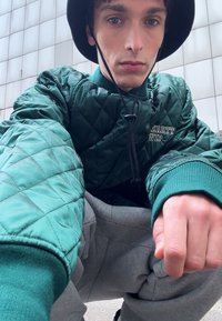 Prešívaná zelená bunda s vysokým golierom a elastickými manžetami, skombinovaná so sivými teplákmi. Model má na hlave čierny klobúk typu bucket.