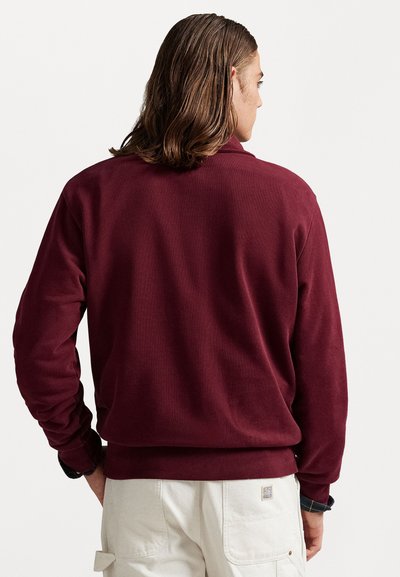 Sweatshirt bordeaux à manches longues, avec des poignets et un ourlet côtelés, et un col rond. Tissu texturé, porté avec un pantalon cargo de couleur claire.