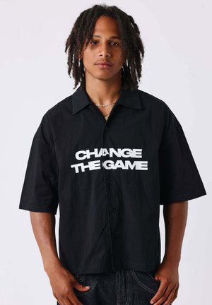 Jeune homme avec des dreadlocks portant une chemise noire à boutons avec un texte blanc "CHANGE THE GAME" à l'avant sur un fond uni.