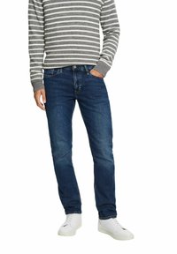 Jean bleu foncé à coupe slim, avec cinq poches et des détails de couture subtils, associé à un pull gris et blanc rayé et des baskets blanches.