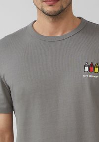 Szara koszulka z krótkim rękawem o ryżowej fakturze, z haftowanymi butelkami przypraw w białym, czerwonym i żółtym kolorze oraz napisem: "POGANIAJMY!"