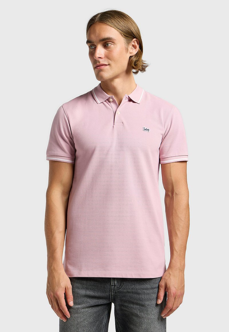 Polo shirt rose clair à manches courtes, avec une patte de boutonnage à deux boutons et un col blanc contrastant. Présente un petit logo bleu sur la poitrine.
