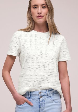 Kvinde med langt blondt hår iført en hvid tekstureret kortærmet top og lyseblå jeans med høj talje og to knapper.