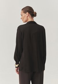 Sort langærmet skjorte med en afslappet pasform, glat tekstur og subtil glans. Har krave og knappede manchetter, stylet med et armbånd med guldaccent.