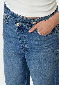 Jeans iz denima v svetlo modri pranja z visokim pasom, z zadrgo, dvema kovinskima gumboma in sprednjim žepom s strganimi robovi.