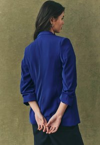 Royal blue blazer with a tailored fit, slightly puffed sleeves, and a smooth texture. Back view shows subtle seam details and a straight hem.