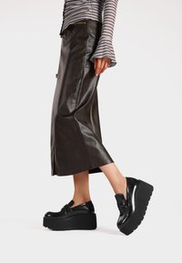 Scarpe platform nere con finitura lucida, caratterizzate da dettagli cuciti e suola spessa. Indossate con una gonna in pelle scura con spacco laterale.