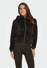 Sort imiteret pels zip-hoodie med hætte, rib-strikkede manchetter og kant. Bæret over mørke rib-strikkede leggings, som præsenterer et struktureret, hyggeligt udseende.