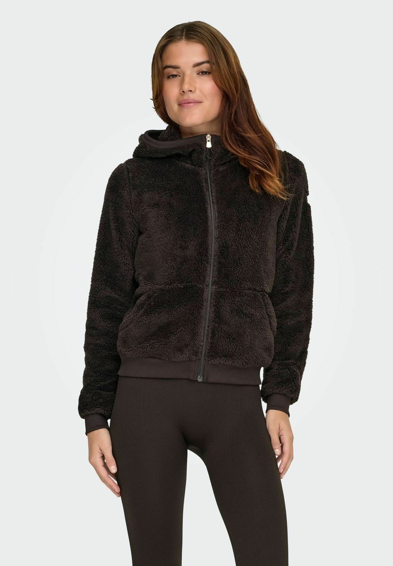 Sort imiteret pels zip-hoodie med hætte, rib-strikkede manchetter og kant. Bæret over mørke rib-strikkede leggings, som præsenterer et struktureret, hyggeligt udseende.