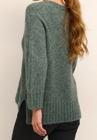 Pull en maille verte avec une coupe décontractée, des poignets côtelés et des fentes sur les côtés, fabriqué à partir d'un matériau doux et texturé, présentant un aspect duveteux.