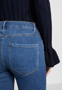 Vaqueros de mezclilla azul con cintura de altura media, con bolsillos traseros y costuras amarillas. Combinados con un suéter azul marino acanalado.