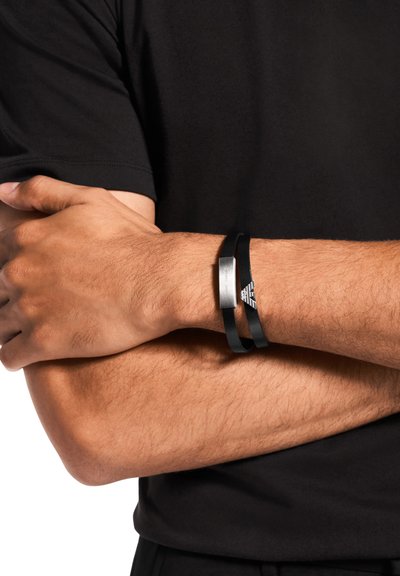 Bracelet noir avec éléments en caoutchouc et métal, présentant un design élégant et un accent métallique gravé, porté sur un poignet avec les bras croisés.