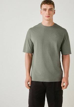 Junger Mann, der ein schlichtes olivgrünes T-Shirt mit kurzen Ärmeln und schwarze Cargo-Hosen trägt, steht vor einem hellen neutralen Hintergrund.