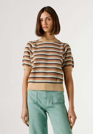 Jonge vrouw met recht bruin haar, gekleed in een gestreepte korte mouw geribbelde trui en lichtgroene broek met hoge taille, tegen een witte achtergrond.