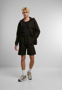 Homme blond portant un sweat à capuche noir zippé, un débardeur noir, un short noir, des chaussettes blanches et des chaussures de course argentées, se tenant devant un fond gris uni.