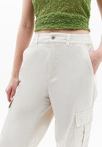 Světle béžové cargo kalhoty s elastickým pasem, bočními kapsami a jemnou texturou. Obsahují detaily prošívání a mají ležérní design.