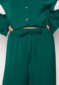 Haut turquoise à boutons avec des manches larges, associé à un pantalon à taille élastique assorti avec un détail de nœud à l'avant dans un tissu lisse.