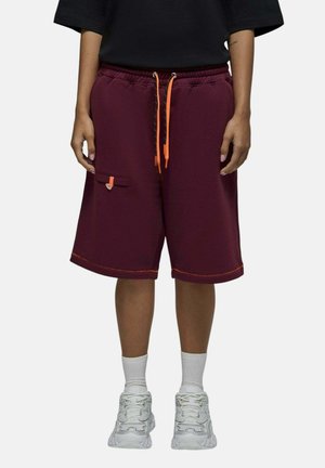Personne portant un short rouge foncé arrivant au genou avec des cordons orange et des baskets blanches, debout devant un fond uni.