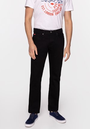 Mężczyzna w czarnych dżinsach, białej koszulce z nadrukiem i niebieskich sznurowanych trampkach stojący na białym tle.