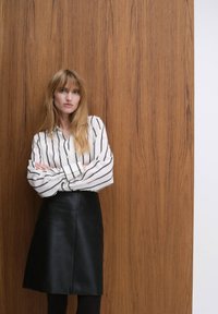 Vit randig skjorta med knappar och svart läderkjol i midjelängd. Bakgrunden är en texturerad träpanel som förstärker outfitens kontrast.
