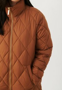 Giacca puffer trapuntata marrone con colletto alto, chiusura con zip e dettagli in oro, e tasche laterali. Tessuto testurizzato con motivi a diamante.