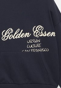Námornícky modrý sveter s bielym textom: "Golden Essen," "URBAN," "KULTÚRA," "SAN FRANCISCO." Obsahuje predné vrecko. Hladká textúra.
