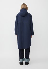 Cappotto lungo blu navy con capuccio aderente, dotato di un design con yoke sulla schiena, tasche profonde e una silhouette rilassata abbinata a jeans scuri con risvolto.