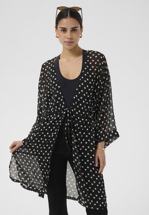 Femme portant un kimono noir transparent à pois sur un top et un pantalon noirs, tenant un côté étendu, lunettes de soleil sur la tête, boucles d'oreilles et bracelet en or.