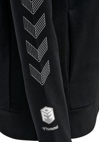 Svart sweatshirt med vita chevron-mönster längs ärmen och en logotyp på cuffen. Tillverkad av mjukt tyg med avslappnad passform.